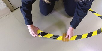 Kuva, jossa henkilö tarttuu kelta-mustanauhaiseen teippiin lattialle
